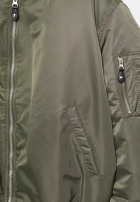 Olive zöld bomberdzseki sima nylonból, bordás mandzsettákkal, teljes cipzárral és ezüst színű fém apróságokkal ellátott oldalsó zsebbel.