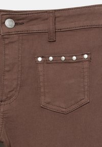 Närbild på bruna denimbyxor som visar en liten framficka med fem silvernitar och en knappstängning vid midjan.
