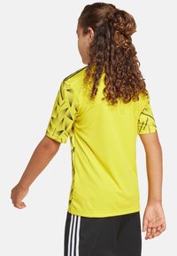 Maglia sportiva gialla brillante con maniche corte, caratterizzata da un motivo geometrico nero lungo le spalle e i lati, realizzata in tessuto leggero.