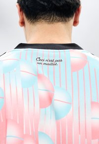 Μικρό μαύρο κείμενο "Ceci n'est pas un maillot" στην πλάτη ενός πουκαμίσου σε παστέλ ροζ και μπλε μοτίβο, που φοριέται από ένα άτομο που φαίνεται από πίσω.