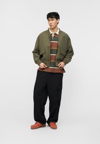 Νεαρός άνδρας που φοράει χακί μπουφάν τύπου μπόμπερ πάνω από μια ριγέ ράγκμπι μπλούζα, χαλαρό μαύρο παντελόνι cargo και καφέ δερμάτινα παπούτσια, στέκεται μπροστά σε ένα μονόχρωμο φόντο.