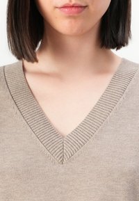 Donna con capelli castano scuro lisci, che indossa un maglione a maglia beige con scollo a V, visibile dalla gola al petto su uno sfondo bianco.