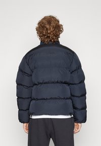 Mornarska puhovka z quiltanim dizajnom, ki ima visok ovratnik, elastične manšete in stranske žepe. Gladek, sijoč material z debelim, toplotno izoliranim videzom.