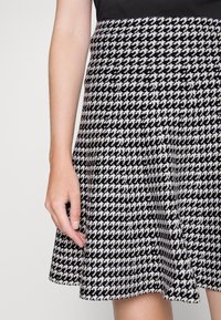 Nainen, jolla on päällään mustavalkoinen houndstooth-kuvioinen hame ja musta toppi, vasen käsi ja käsivarsi rentona sivulla.