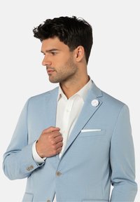 Veste de costume bleu clair avec une finition texturée, revers en pointe, fermeture à un bouton et un pochette blanche. Accentuée d'une épingle à revers blanche.