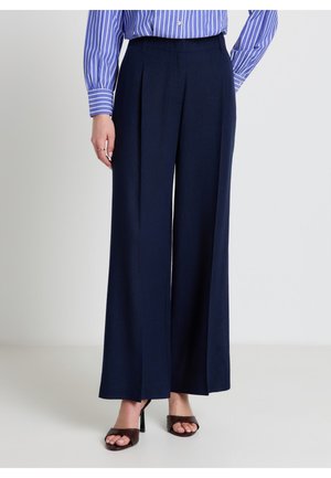 Femme portant un pantalon large bleu marine, une chemise rayée bleu et blanc, et des sandales noires à talons hauts, debout devant un fond uni.