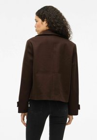 Chaqueta de mezcla de lana marrón con una silueta estructurada, grandes solapas y puños abotonados, combinada con unos jeans negros ajustados.