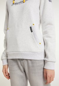 Světle šedá mikina s žlutými šňůrkami a černým textem na přední kapse, nošena s vybledlými šedými džíny, ruka uvolněně stojí po boku.