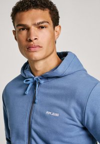 Zils kapucis, kas aizveras ar rāvējslēdzēju, izgatavots no mīksta auduma, ar regulējamu kapuci un izšūtu "PEPE JEANS" logotipu uz krūtīm.