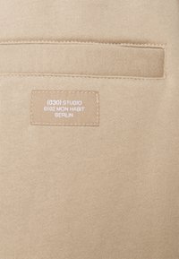 Béžová látka s hladkým povrchem má v centru přišitou obdélníkovou štítku, na níž je napsáno "(030) STUDIO 0102 MON HABIT BERLIN."