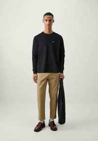 Black long-sleeve t-shirt with a round neck, beige trousers, brown shoes, and a navy blazer draped over the left arm. Minimalist branding.