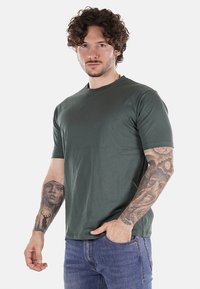 T-shirt verde in cotone con maniche corte, collo rotondo e vestibilità rilassata. Presenta braccia tatuate visibili e jeans in denim blu scuro.