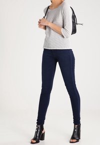 Maglietta a maniche lunghe grigia a righe, jeans skinny scuri e stivaletti con tacco aperti neri. Il modello porta uno zaino nero su una spalla.