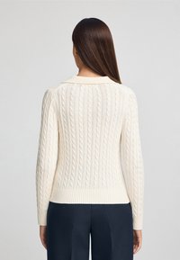 Cremefarbener Kabelstrickpullover mit geripptem Saum und Kragen, langen Ärmeln und einem strukturierten Muster auf der Rückseite.