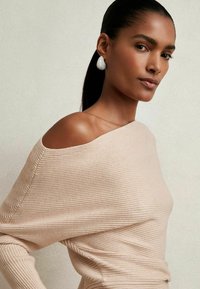 Helles beige, geripptes Oberteil mit einem schulterfreien Design. Das Modell hat eine glatte Textur und eine moderne Silhouette. Große weiße Ohrringe betonen den Look.