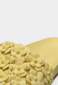 Chaussure jaune sans lacets avec un motif floral texturé, présentant de petites pétales de fleurs en relief sur le devant et une semelle intérieure lisse assortie.