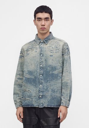 Jeune homme portant une chemise en jean bleu clair usée et boutonnée ainsi qu'un pantalon noir, debout devant un fond blanc uni.