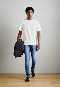 T-shirt bianco in cotone, jeans blu, scarpe da ginnastica nere. Tiene in mano un giubbotto imbottito nero. Design semplice, texture liscia, colori neutri.