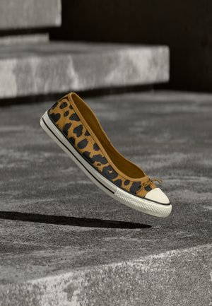 Λευκό sneaker με σχέδιο λεοπάρ, με λευκή λαστιχένια μύτη και σόλα, αιωρείται πάνω από γκρι τσιμεντένια επιφάνεια με σκαλοπάτια στο βάθος.