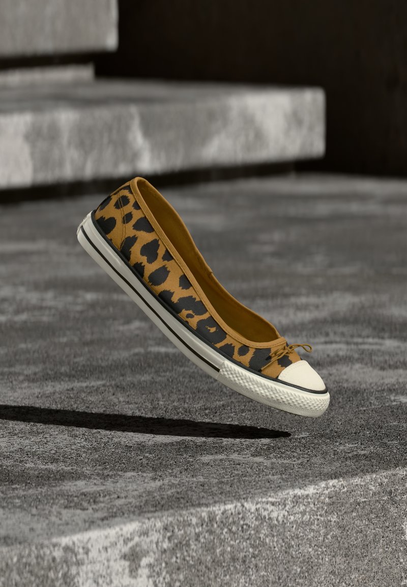 Λευκό sneaker με σχέδιο λεοπάρ, με λευκή λαστιχένια μύτη και σόλα, αιωρείται πάνω από γκρι τσιμεντένια επιφάνεια με σκαλοπάτια στο βάθος.