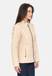 Chaqueta acolchada beige con cuello alzado, bolsillos laterales con cremallera y un patrón diagonal. Textura de tela suave con detalles de cremallera en oscuro.