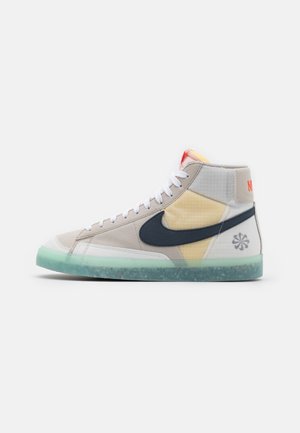 Wysokie sneakersy z beżowymi, żółtymi i szarymi panelami, czarnym logo Nike, białymi sznurowadłami i przezroczystą zieloną podeszwą z plamkami.