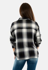 Camisa de franela a cuadros en blanco y negro con un ajuste relajado, mangas enrolladas y sutilezas decorativas, que presenta una parte trasera más larga para mayor cobertura.