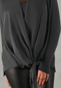 Top negro de wrap, hecho de una tela ligera y suave, con un cierre de lazo en el frente y mangas largas, combinado con pantalones negros ajustados.