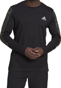 Muž v černém tričku Adidas s dlouhými rukávy a tmavozelenými pruhy na rukávech, ruce volně spojené před sebou, obličej není viditelný.