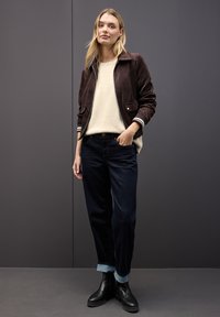 Veste bomber en daim marron avec des détails de manches à rayures blanches, pull crème, jean en denim foncé avec les poignets retroussés, et bottines noires.