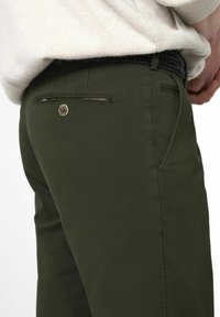 Olivgrüne Hosen mit einer glatten Textur, ausgestattet mit einem goldenen Knopf und einem Detail an der Gesäßtasche. Begleitet von einem gemusterten, gewebten schwarzen Gürtel.
