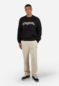Black knit sweater with fuzzy "Plegador" lettering. Beige pleated trousers and black sneakers with white soles complete the outfit.