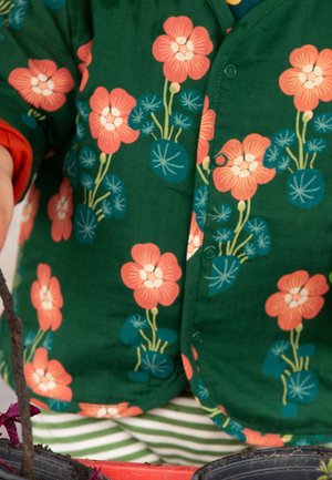 Green jacket with orange and blue floral pattern worn over green and white striped pants, partial view of a person holding a plant.