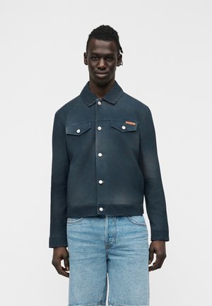 Man draagt een donkerblauwe jas met knopen, kraag en borstzakken, gecombineerd met lichtblauwe denim shorts, staand tegen een effen achtergrond.