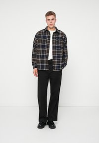 Camicia di flanella a quadri nei colori blu scuro, marrone e crema, con due tasche sul petto. Indossata su una maglietta bianca e abbinata a pantaloni neri.