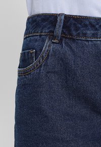 Tmavě modré džínové kalhoty s klasickým designem pěti kapes, zesíleným prošíváním a páskem zakončeným kovovým zapínáním.