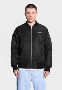 Czarna kurtka bombowa wykonana z gładkiego materiału, posiada zamek błyskawiczny z przodu, ściągacze przy mankietach, boczne kieszenie i detal z logo na klatce piersiowej.