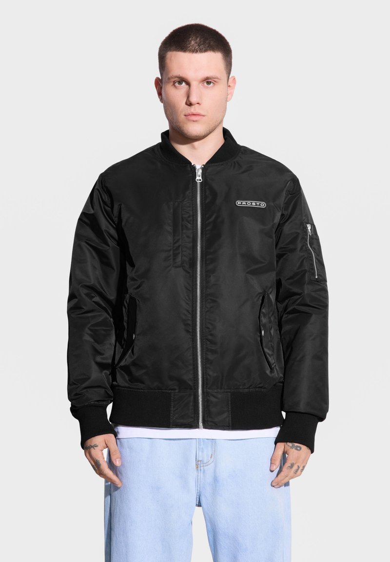 Czarna kurtka bombowa wykonana z gładkiego materiału, posiada zamek błyskawiczny z przodu, ściągacze przy mankietach, boczne kieszenie i detal z logo na klatce piersiowej.