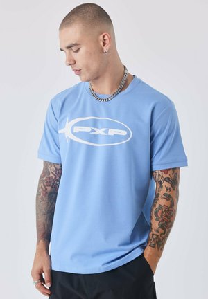 Blauwe T-shirt met korte mouwen heeft een wit logo op de borst. De stof is glad met een ontspannen pasvorm. Het model heeft tatoeages en accessoires.
