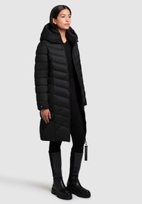 Kvinde iført en lang sort quiltet pufferjakke med høj krave, parret med sorte leggings og høje sorte støvler.