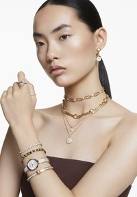 Joyería de oro exhibida en un modelo, que presenta collares en capas, anillos y un reloj con carátula blanca, acentuado con rhinestones y variaciones de textura.