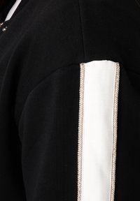 Vêtement noir avec un panneau blanc et une bordure tressée le long de l'épaule. Le tissu semble doux avec une texture lisse.