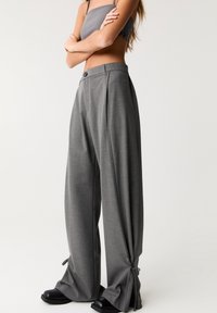 Pantalons plissés gris à taille haute, avec des détails de noeud sur les côtés et une coupe large. Portés avec un top cropped gris assorti et des chaussures noires épaisses.