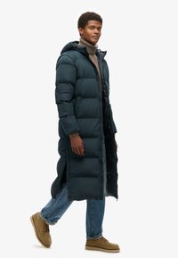 Donkergroene, lange pufferjas met een capuchon, voorzien van een rits aan de voorkant en zijsplitten. Draag het over een lichte trui en een blauwe spijkerbroek, gecombineerd met beige schoenen.