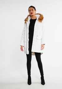 Witte parka jas met een bontgevoerd capuchon, uitgevoerd met gouden hardware, ritssluiting en voorzakken. Het model draagt zwarte leggings en laarzen.