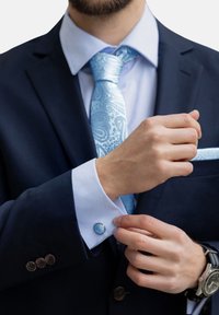 Jūrų laivyno kostiumas su šviesiai mėlyna marškinėlių ir paisley rašto kaklaraiščiu. Rankogalių segtukai ant baltos rankovės, ranka reguliuojanti rankovę, matomas laikrodis.