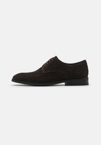 Zapato de vestir de ante marrón oscuro con una textura suave, punta redondeada y un sistema de cordones con cinco ojales. Diseño elegante sobre una suela baja y plana.