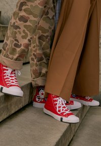 Sneaker alti rossi con lacci e punte bianche, abbinati a pantaloni larghi mimetici e marroni, posizionati su un gradino di pietra.