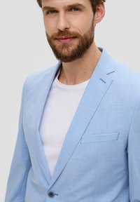 Lichtblauwe blazer van textuurstof, met een opstaande kraag, enkele knoopsluiting en een borstzak. Draag over een witte t-shirt.