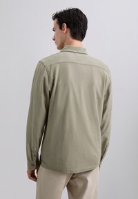 Camisa de botón de manga larga en tejido de color verde oliva claro, con cuello, botones en los puños y un dobladillo en curva. Textura suave, diseño casual.